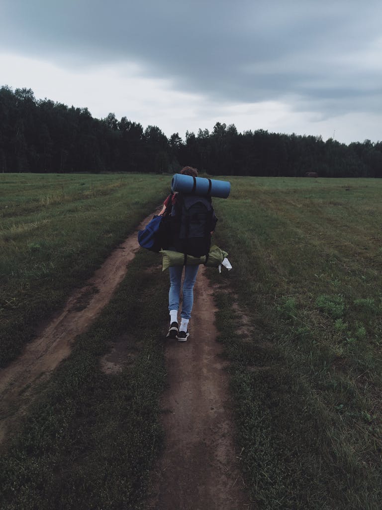 A solo backpacker walking along an unpaved road in a lush green landscape.