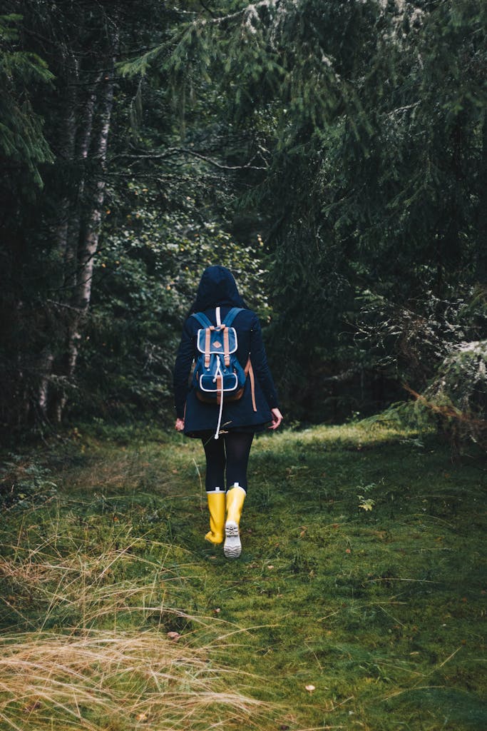 Back view of unrecognizable hiker in outerwear and yellow rubber boots walking in forest in summer day